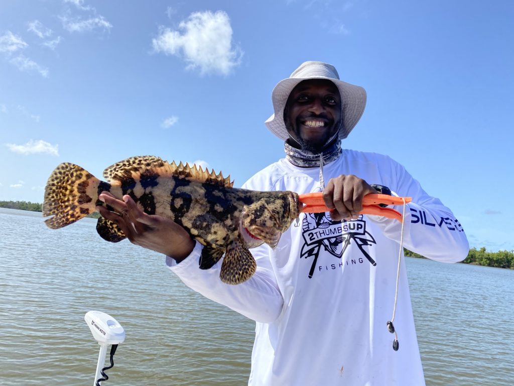 A smiling Captain of 2 Thumbs Up Charters holding Grouper on a boat 
