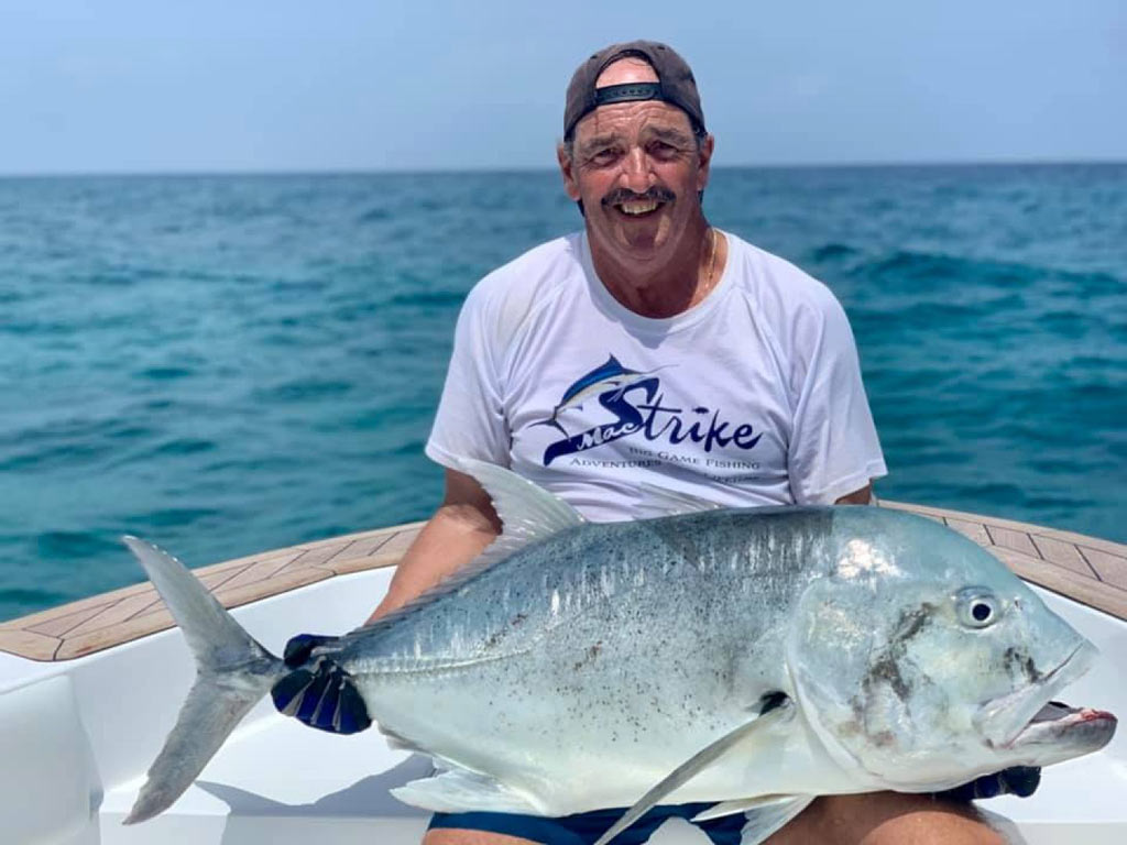 An angler on a fishing charter, wearing a backwards baseball cap, holding a large Giant Trevally, with crystal clear ocean waters behind him