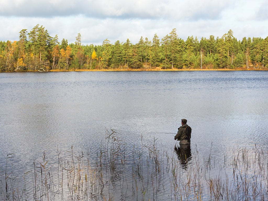 An angler standing in a lake, holding a fishing rod on a cloudy day, with a tree-lined shore in the distance.