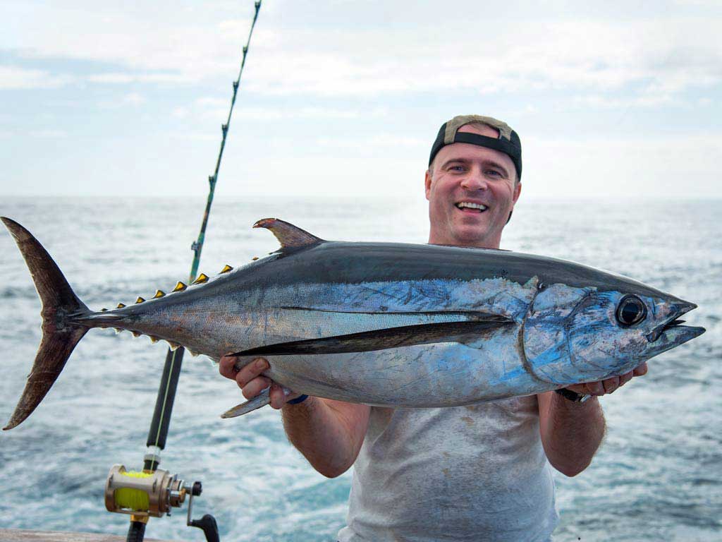 A happy fisherman smiling and holding a large albacore tuna with both hands in a boat.
