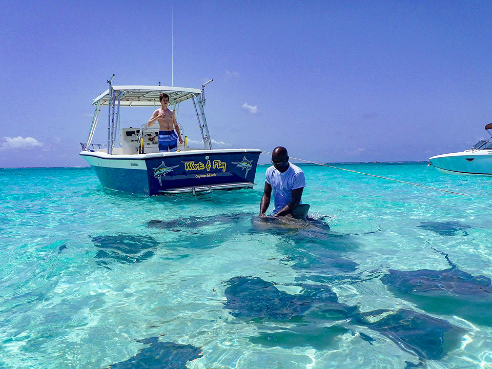 Two anglers in the Cayman Islands, one standing in the water holding a Ray, and the other standing on a boat on a clear sunny day.