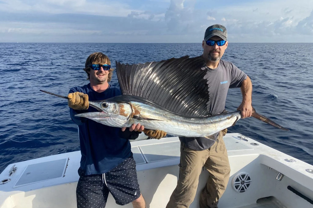 Two men stand aboard a charter boat holding a Sailfish, a popular kite fishing target