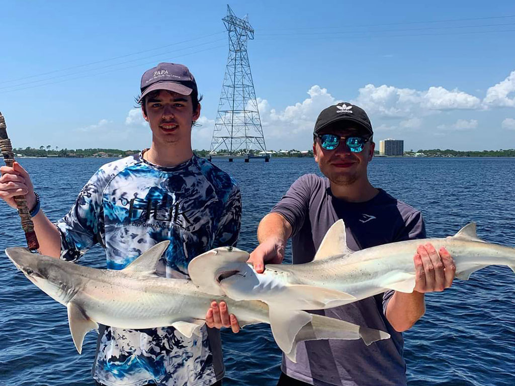 Dos pescadores sostienen un tiburón cada uno con un pilón detrás de ellos en el agua