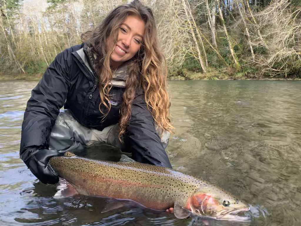 A cute woman smiling and holding a Rainbow Trout with both her hands slightly touching water