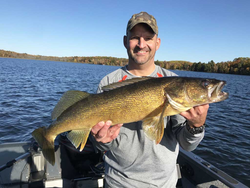 A proud angler in a camo hat and a sweatshirt, standing on a boat, holding a Walleye he caught with both his hands