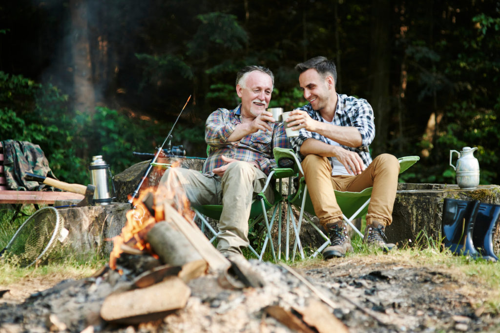 Two fishermen drinking coffee by a bonfire while camping.
