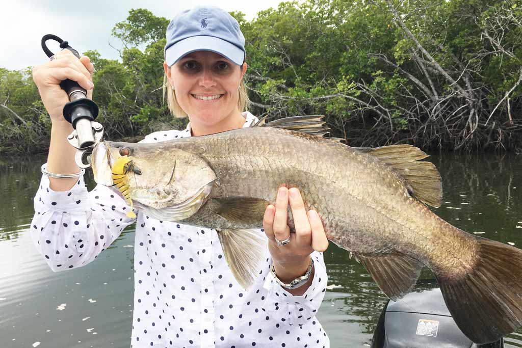 A smiling fisherwoman holding a Barramundi with mangroves in the background