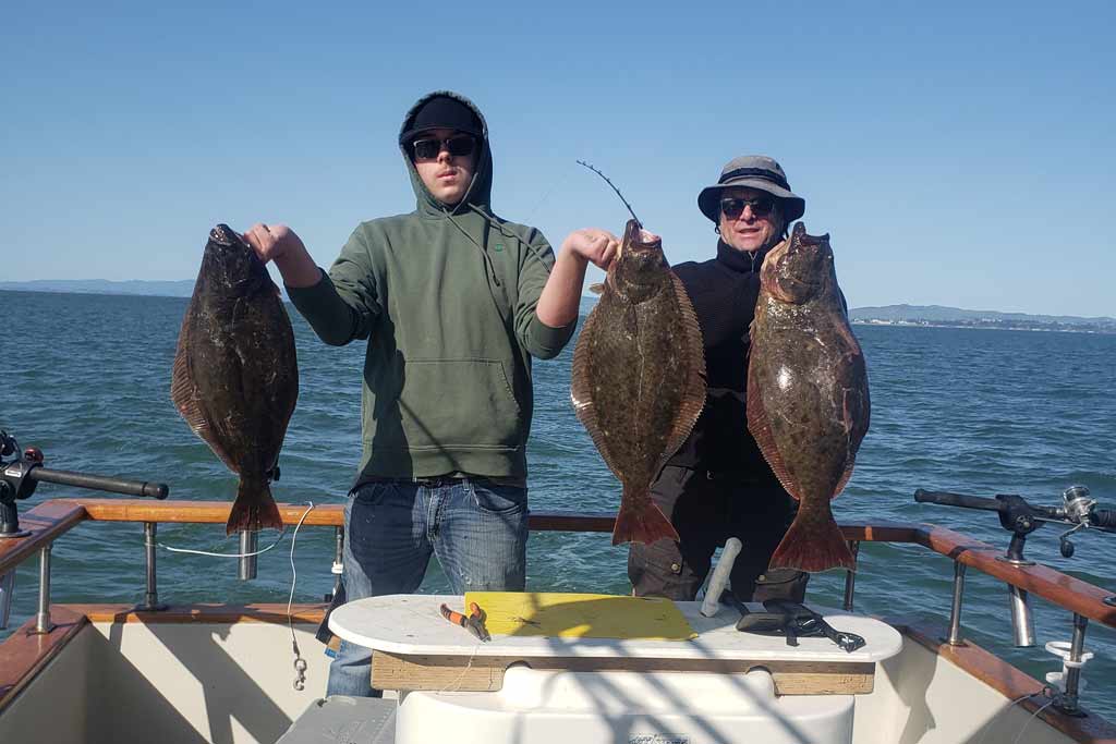 Two anglers standing on a boat, holding three Halibut, blue skies and water in the background