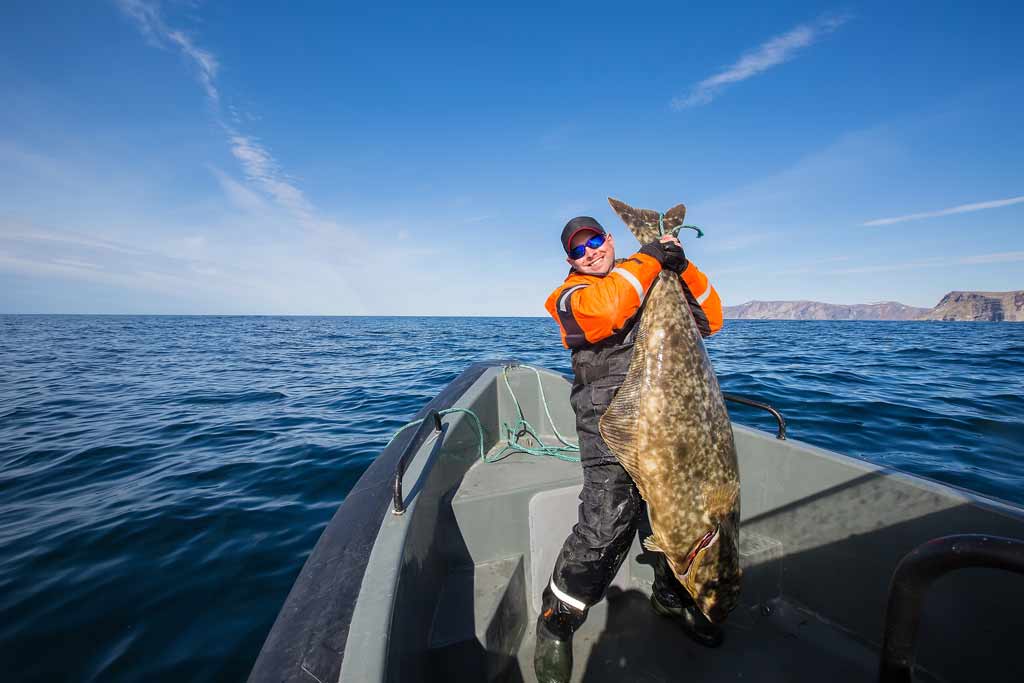 A fisherman standing on a boat, holding a big Pacific Halibut, blue skies and water in the background