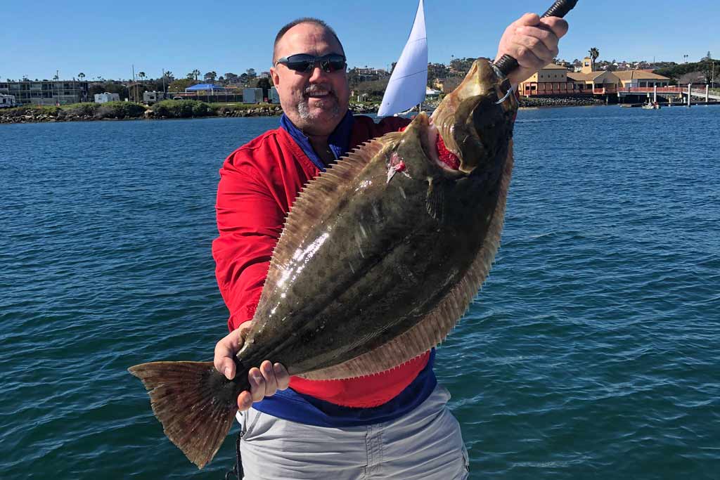 A smiling fisherman in sunglasses, standing on a boat, holding a Halibut