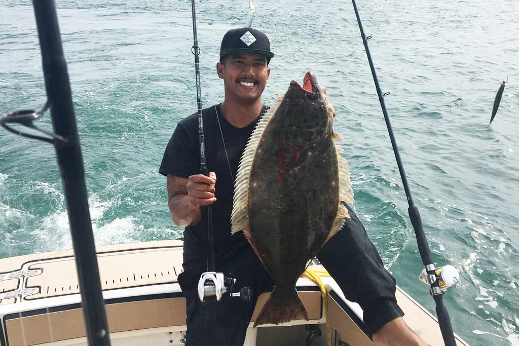 A smiling angler in a cap sitting on the side of the boat, holding a Halibut, fishing rods around him