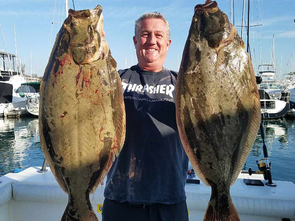 A smiling fisherman standing on a dock, holding two big California Halibut