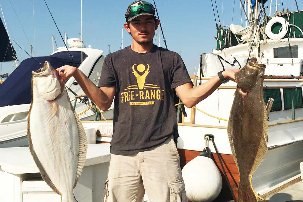 A man standing on a dock, holding two Halibut, fishing boats in the background