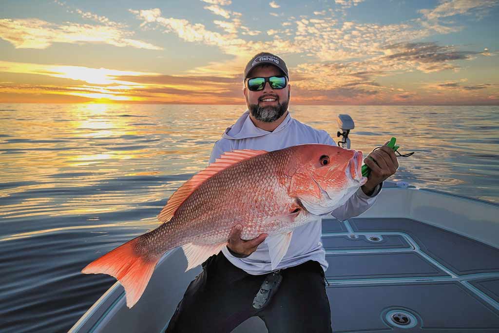 A fisherman in a cap and sunglasses, sitting on a boat, holding a big Red Snapper, sunset in the background