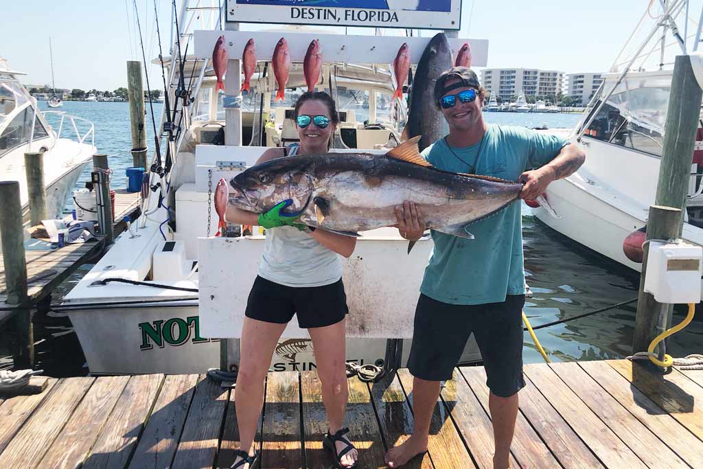 A man and a woman standing on a dock, holding an Amberjack fish, with a boat behind them