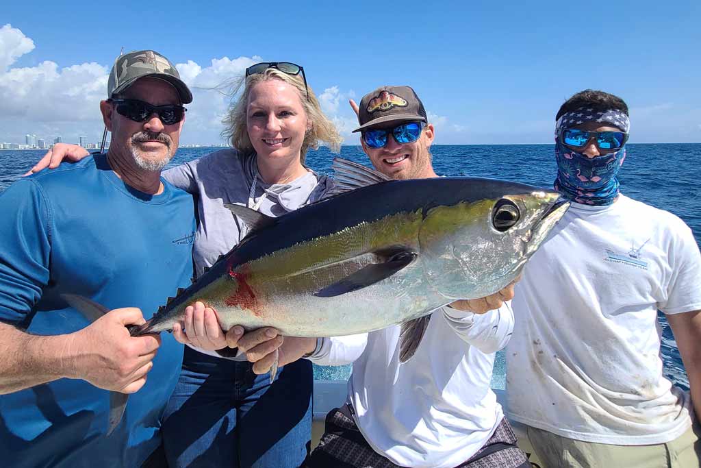 Three men and a woman standing on a boat, holding a Blackfin Tuna with the water behind them and a cityscape in the distance