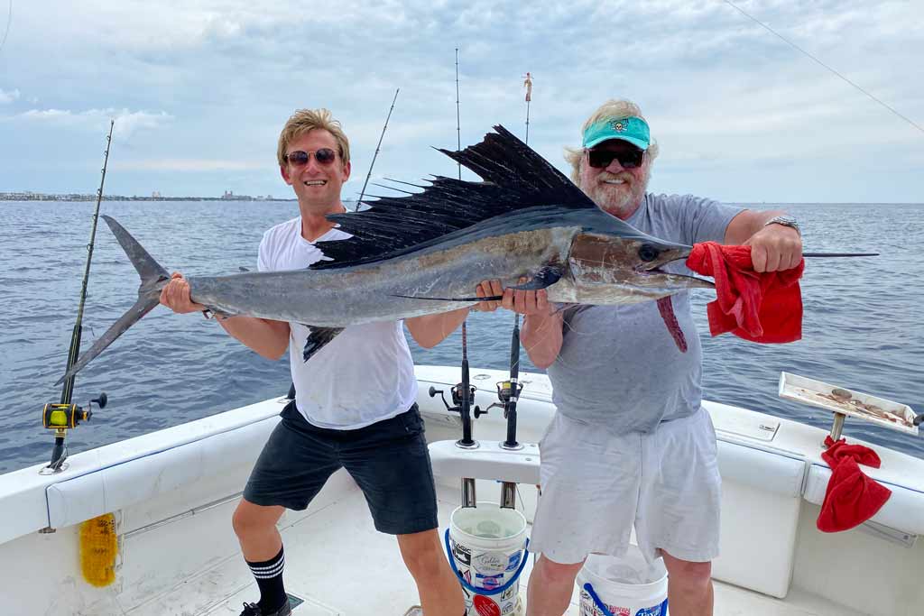 One middle-aged man and flipside younger one holding a Sailfish they just caught, while standing on a wend with water overdue them and land in the distance