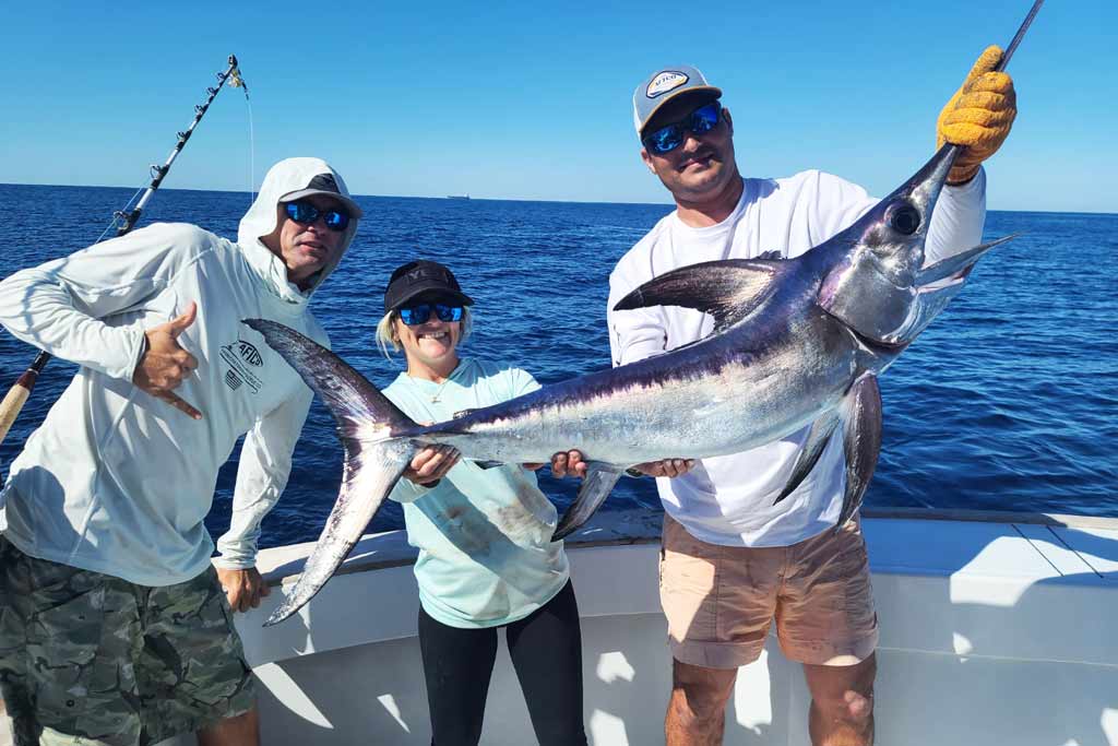 A woman and two men holding their catch of a large Swordfish on a boat