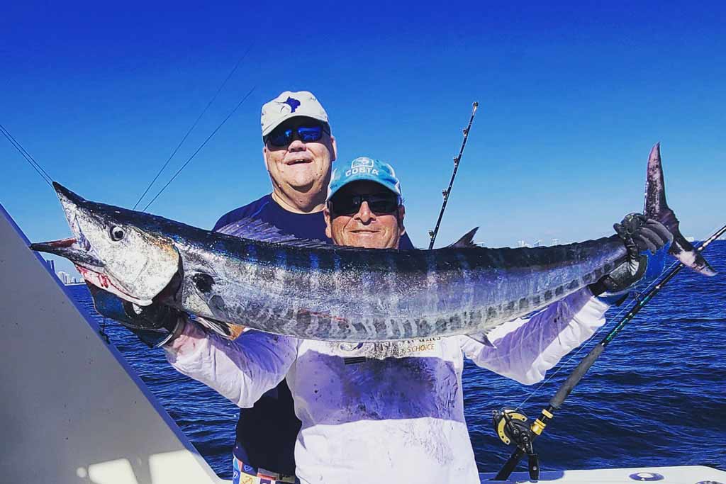 Two anglers on a boat, holding a big Wahoo, with blue water and clear skies in the background
