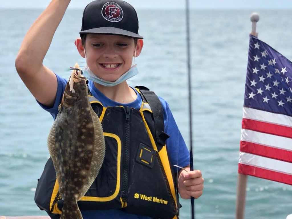 A young boy in a baseball cap, proudly looking at his Fluke catch next to an American flag on a flagpole on a boat