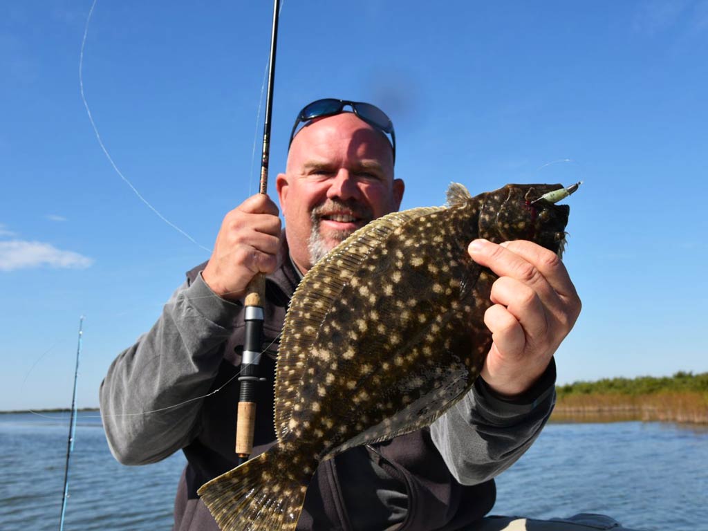 An angler holding a Flounder in one hand and a fishing rod in other hand while on a boat.