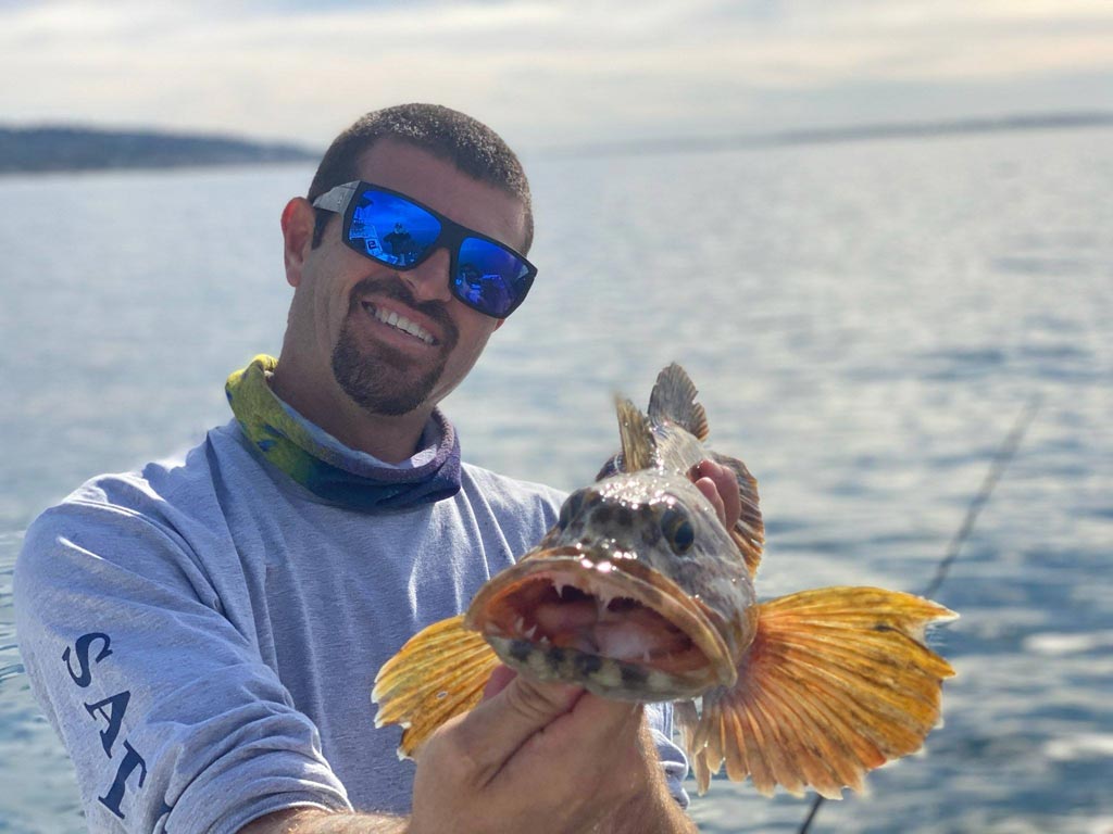 An angler holding a Lingcod towards the camera with its open mouth visible.