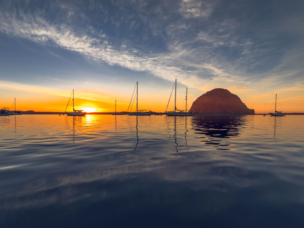 The sunset in Morro Bay with several sailboats visible in the water and Morro Rock in the distance.