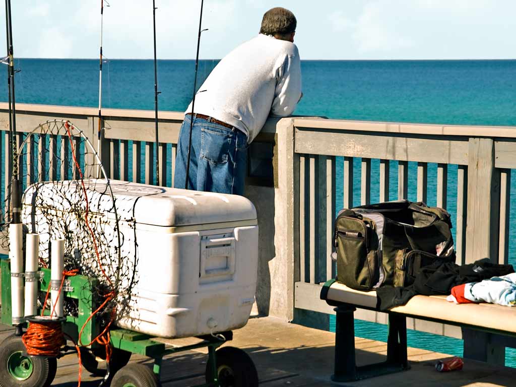 A view of fishing equipment like rods and cooler behind an angler looking out, preparing to fish from a pier