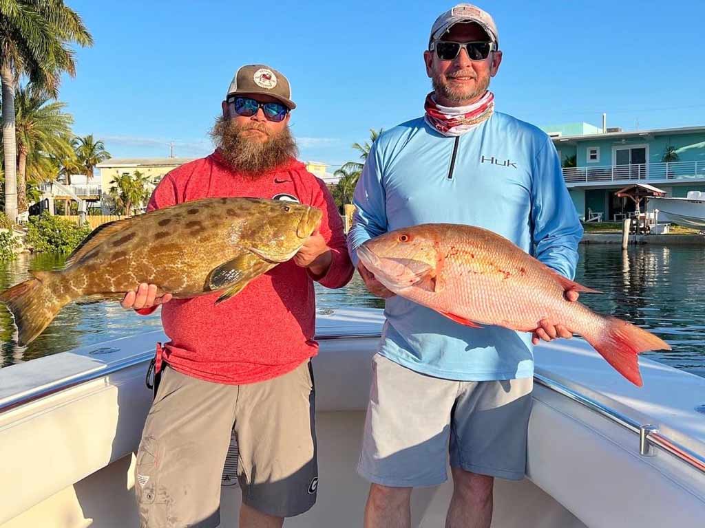 Two anglers standing on a boat and holding a Grouper and Snapper each, caught while fishing in Islamorada