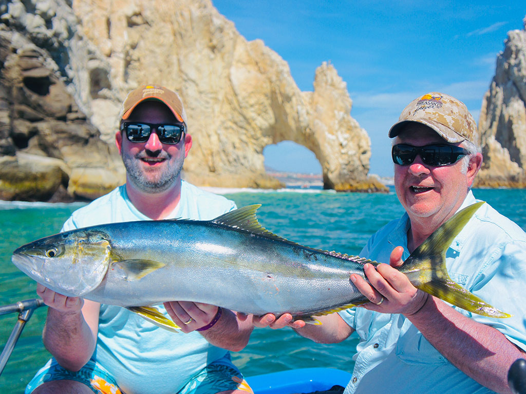 Two anglers hold a Yellowtail Amberjack in front of Cabo San Lucas's famous arch on the water