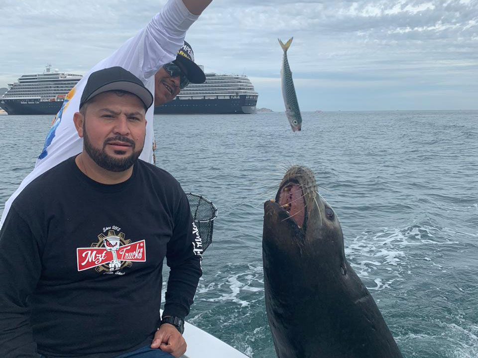 An angler poses on a boat next to a Sea Lion, while someone behind him dangles a fish for the Sea Lion to eat