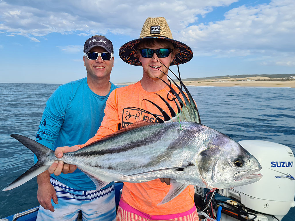 A young anger holds a large Roosterfish with an older person behind them in the shallow waters of Cabo San Lucas