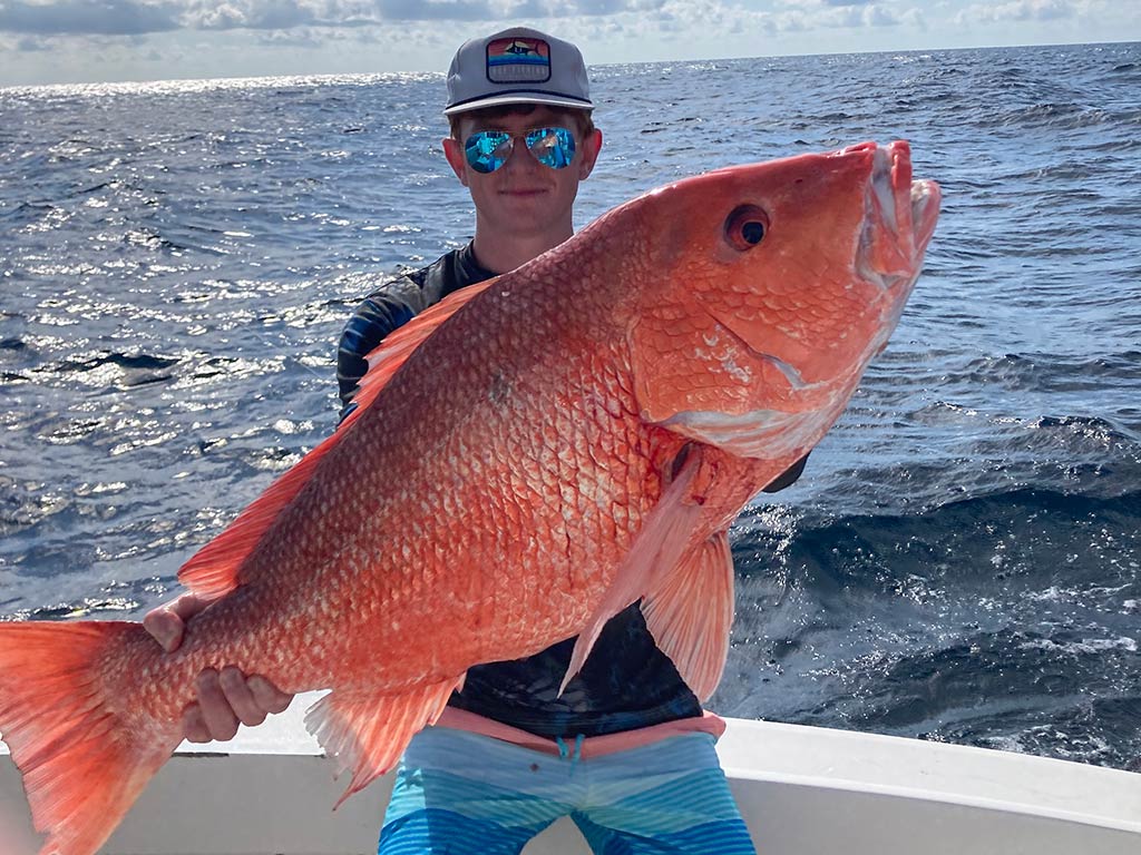 An angler sitting on the side of a boat holding a massive Red Snapper he just caught.