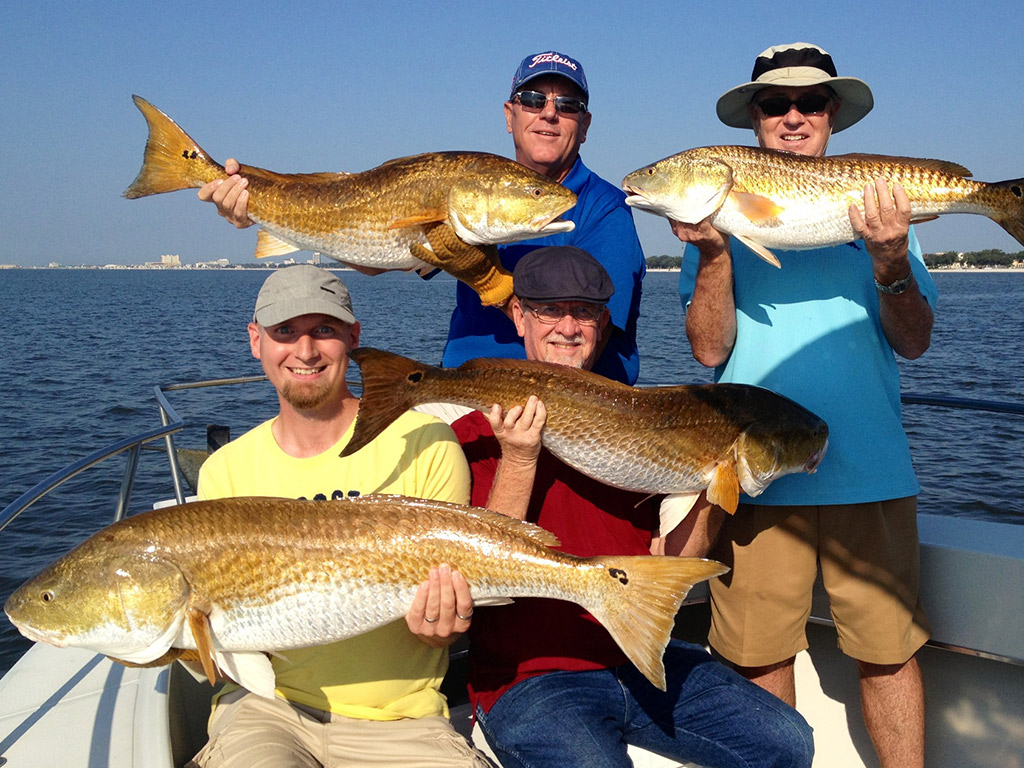 A group of four friends on a fishing charter showing Redfish they caught on their trip out of Gulfport, MS, with the water behind them and land in the distance.