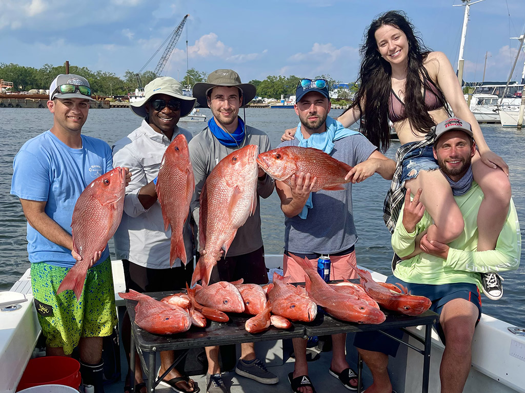 A group of five men and one woman on a man's shoulders standing on the deck of a boat holding limits of Red Snapper they caught while fishing out of Pensacola.
