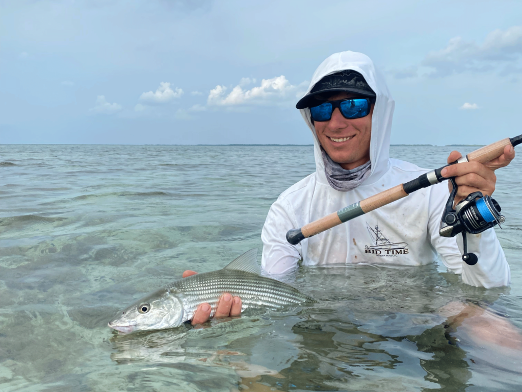 A smiling fly angler sitting in the water in the with a rod in one hand and Bonefish in the other.