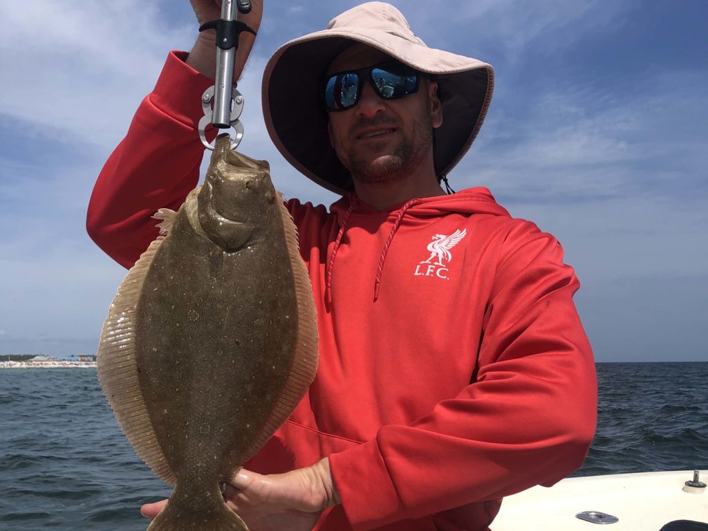 A happy angler in a red hoodie and a light-colored hat holding a freshly-caught Flounder in Orange Beach, Alabama.
