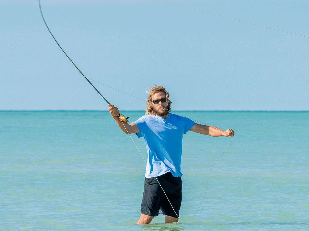 An angler wearing sunglasses casting his fly rod with the flats behind him.