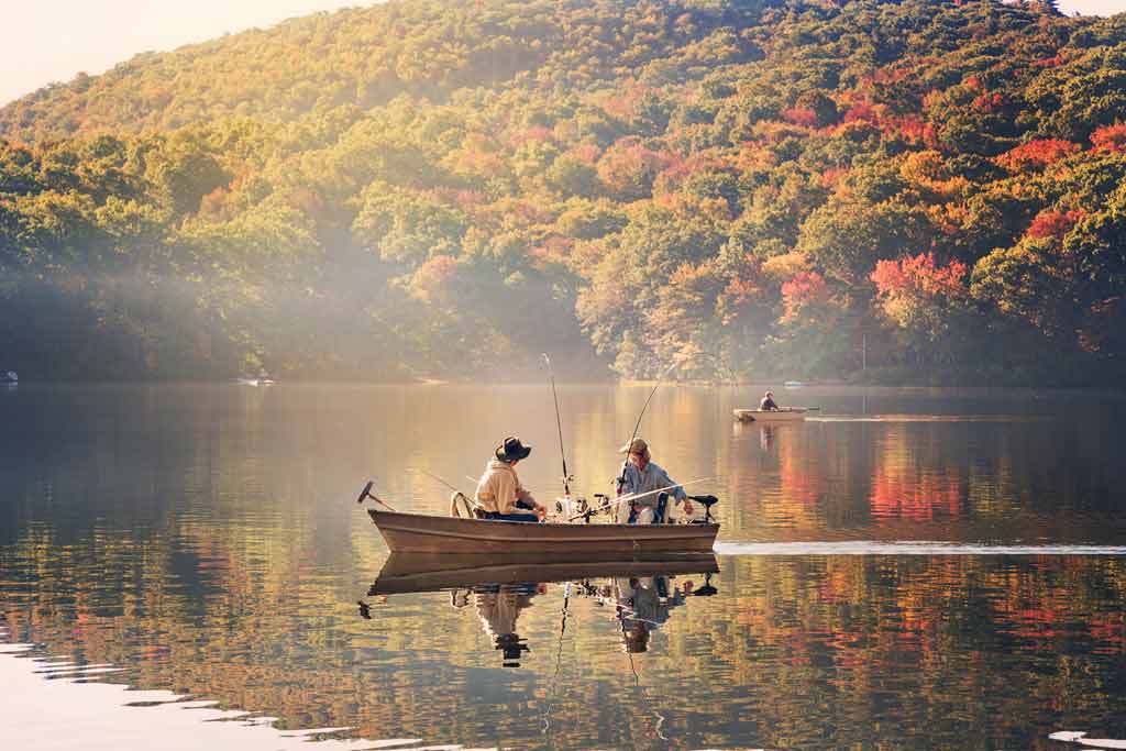 Two angers sitting on a boat, fishing with sunshine and fall foliage in the background
