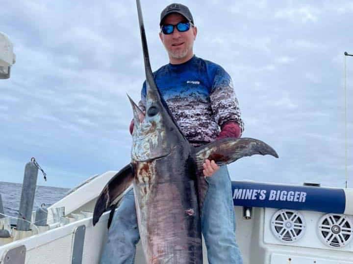 An angler with a hat and sunglasses on standing on a boat in New Jersey and holding a freshly-caught Swordfish.