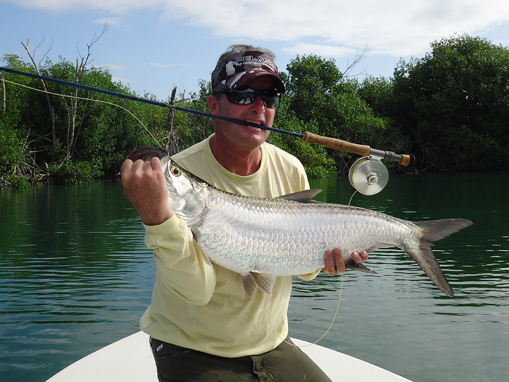 Angler holds a fly rod between his teeth and a Tarpon he just caught somewhere near Cancun in his hands.