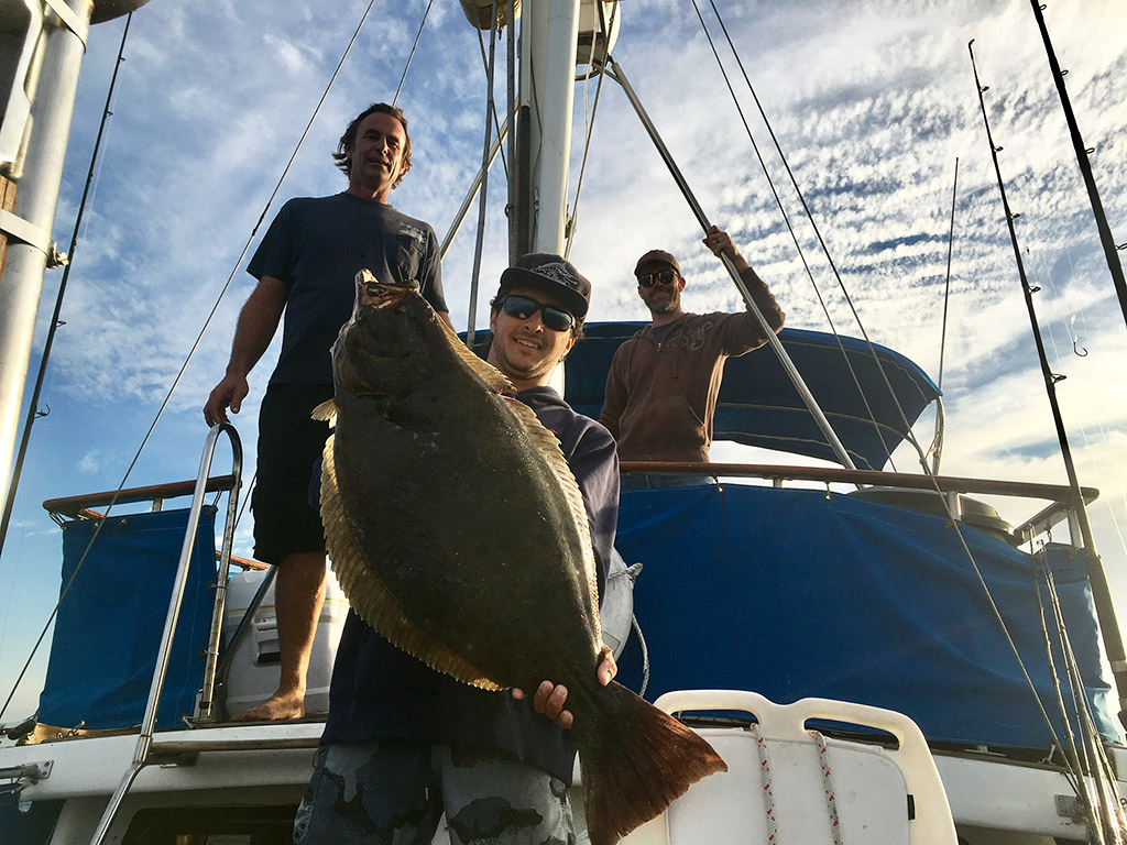 Three anglers stand on a boat, one of them holds a Halibut he just caught on a deep sea trip.