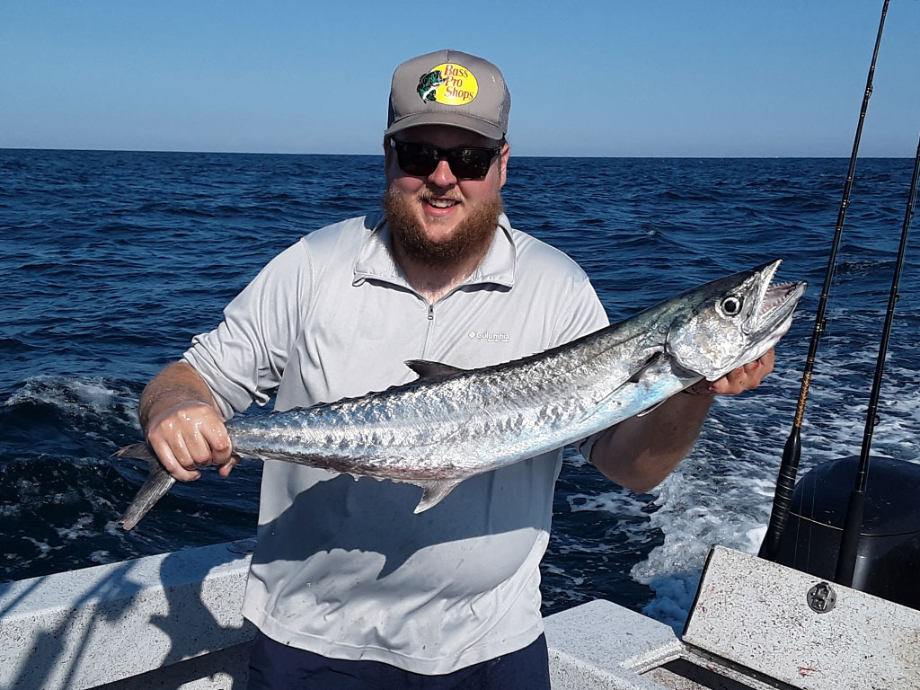 A smiling angler stands on a boat holding a Kingfish he just caught