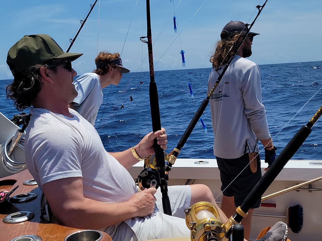 Three anglers on a boat during an offshore trip to the Gulf Stream, with one sitting in a fighting chair holding two rods.