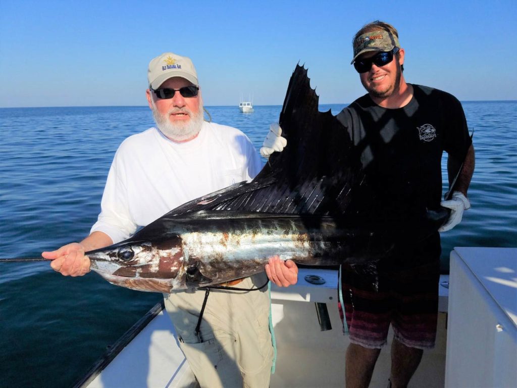 A picture showing two anglers on a charter fishing boat, both holding a freshly caught Sailfish with another boat and offshore waters behind them, Cape Canaveral, Florida