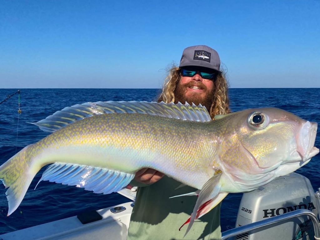 A picture showing a happy angler wearing glasses and a hat, standing on a charter fishing boat with a freshly caught Tilefish on a sunny day in Cape Canaveral, Florida