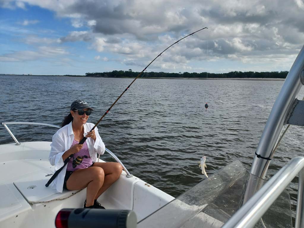 A female angler sitting on a boat and holding a rod with a small fish hooked at the end it.