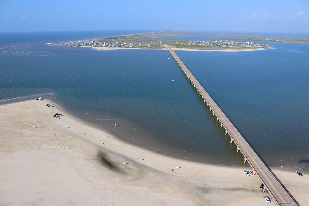 An aerial view of the San Luis Pass Toll Bridge on a sunny day with some cars parked up on the beach