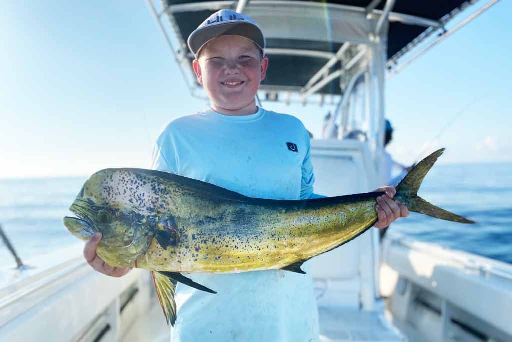 A smiling boy in a cap standing on a fishing boat, holding a Mahi Mahi on a sunny day