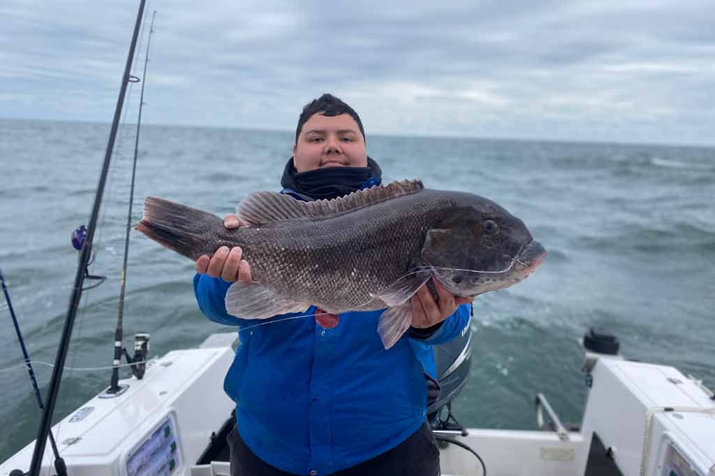 A smiling young man holding a Tautog while standing on a boat, with the water and cloudy skies in the background